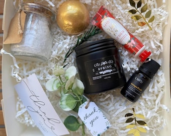 May include: A gift box filled with self-care items. Includes a jar of bath salts, a gold bath bomb, a black candle, lip balm, essential oil, and decorative accents. The box also contains two "Thank You" tags.