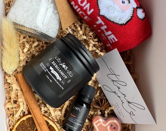 May include: A gift box filled with self-care items. Includes a black candle, a jar of bath salts, a bottle of rose essential oil, a gingerbread man bath bomb, a pair of red socks with Santa, a dried orange slice, cinnamon sticks, and a thank you card.