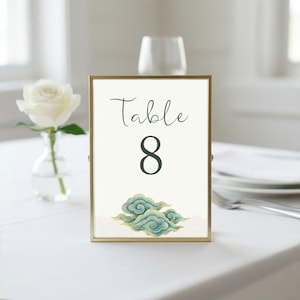 May include: A framed table number card with the word "Table" in script and the number "8" in a dark teal color. The card has a gold frame and features a teal and gold cloud design at the bottom. A white rose in a vase is in the background.