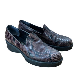 May include: A pair of brown and black snakeskin-patterned slip-on shoes with a low wedge heel. The shoes have a rounded toe and a black sole. The shoes are made of a textured material.