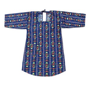 May include: A blue tunic featuring a repeating vertical pattern of colorful floral motifs in red, green, and yellow. The garment has long sleeves, a square neckline with a small tie closure at the center, and a straight hemline that curves slightly.