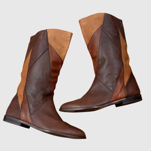 May include: A pair of mid-calf boots featuring a multi-tonal patchwork design in shades of dark brown and tan. The boots have a flat sole, a rounded toe, and a vertical seam construction that creates geometric panels across the entire surface.