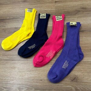May include: Four pairs of crew socks in vibrant colors: yellow, navy blue, hot pink, and purple. Each sock has a tag at the top and text near the foot. The socks are arranged on a wooden surface.