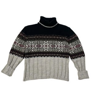May include: A turtleneck sweater with a fair isle pattern. The top is black, transitioning to a pattern of white, burgundy, and olive green geometric shapes. The lower portion is a light gray with ribbed texture, and the sleeves have folded cuffs.