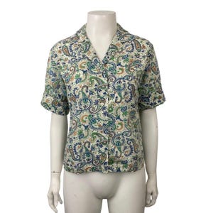 May include: Short-sleeved button-down shirt with a paisley pattern in shades of blue, green, and brown on an off-white background. The shirt has a classic collar and is displayed on a mannequin.