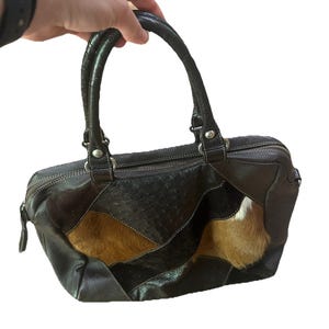 May include: A dark brown leather handbag with a patchwork design. The bag features a top handle, a zipper closure, and a mix of textures, including smooth leather and textured panels. The bag is held by a hand.