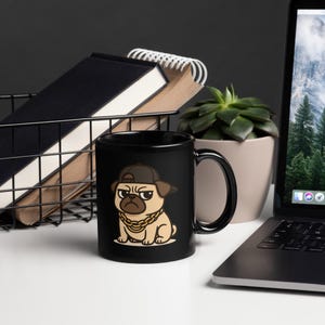 May include: Black ceramic mug featuring a cartoon pug wearing a brown baseball cap and gold chain. The pug has a stern expression. The mug is on a white surface with a laptop, plant, and wire basket in the background.