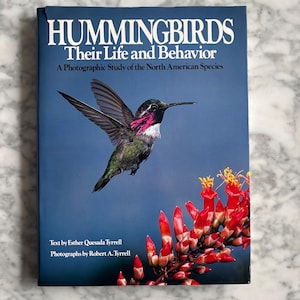 Può includere: Un libro intitolato "HUMMINGBIRDS Their Life and Behavior" con una fotografia di un colibrì in volo. La copertina blu presenta testo bianco e un'immagine dell'uccello e fiori rossi.