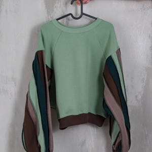 Peut inclure: Un sweat-shirt vert sauge avec des bordures côtelées marron et des manches rayées multicolores. Les manches présentent des rayures verticales dans les tons marron, vert et mauve. Le vêtement est suspendu à un cintre noir.