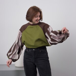 Könnte beinhalten: Ein einzigartiges Sweatshirt mit Farbblöcken in Olivgrün und Braun. Die Ärmel haben ein Streifenmuster in Braun- und Cremetönen. Die Person im Bild trägt eine schwarze Jeans.
