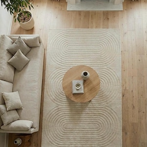 Neutral modern area rug hand tufted from premium wool in 8x10 dimension. The abstract curved motif creates movement while staying subtle and elegant. Photographed in a bright living space with beige sofa, wooden table, and fireplace. Excellent for homeowners seeking contemporary, Scandinavian, or organic modern decor with comfort and high end appeal.