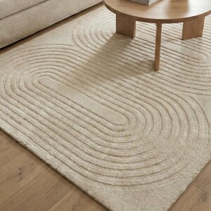 Scandinavian style hand tufted rug in cream and beige, size 8x10, with dimensional abstract pattern. Positioned in a sunlit room near a fireplace and indoor plant, the rug highlights clean lines and cozy living. Soft wool construction ensures comfort while the neutral color matches many decor themes from minimalist to contemporary homes.