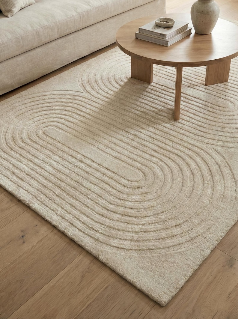 Scandinavian style hand tufted rug in cream and beige, size 8x10, with dimensional abstract pattern. Positioned in a sunlit room near a fireplace and indoor plant, the rug highlights clean lines and cozy living. Soft wool construction ensures comfort while the neutral color matches many decor themes from minimalist to contemporary homes.