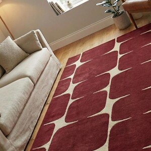 Luxury hand tufted geometric area rug in burgundy and ivory colors showcased in a minimal modern living room interior. This handmade wool rug features a unique abstract pattern that complements neutral furniture, wooden flooring, and indoor plants. Perfect for modern home decor, this soft and durable rug adds comfort, warmth, and visual interest to living rooms, bedrooms, apartments, and contemporary spaces.