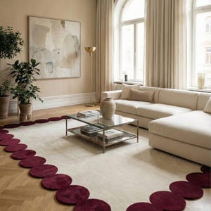 Burgundy scalloped hand tufted rug size 8x10 placed in a bright modern living room with cream sectional sofa, glass coffee table and tall arched windows. The soft ivory center is framed by rich wine red circular edges creating a bold statement look while keeping the interior minimal, warm and luxurious. Plush wool texture makes it ideal for cozy seating areas and contemporary homes.