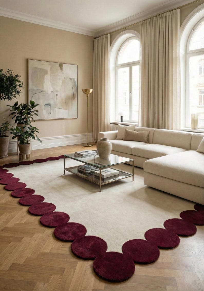 Burgundy scalloped hand tufted rug size 8x10 placed in a bright modern living room with cream sectional sofa, glass coffee table and tall arched windows. The soft ivory center is framed by rich wine red circular edges creating a bold statement look while keeping the interior minimal, warm and luxurious. Plush wool texture makes it ideal for cozy seating areas and contemporary homes.