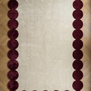 Hand tufted wool rug size 6x9 designed with a unique burgundy bubble border and subtle ivory base. Positioned in a stylish lounge near plants and modern furniture, the plush surface enhances warmth and texture. Ideal for homeowners seeking a fashionable yet timeless living room upgrade.