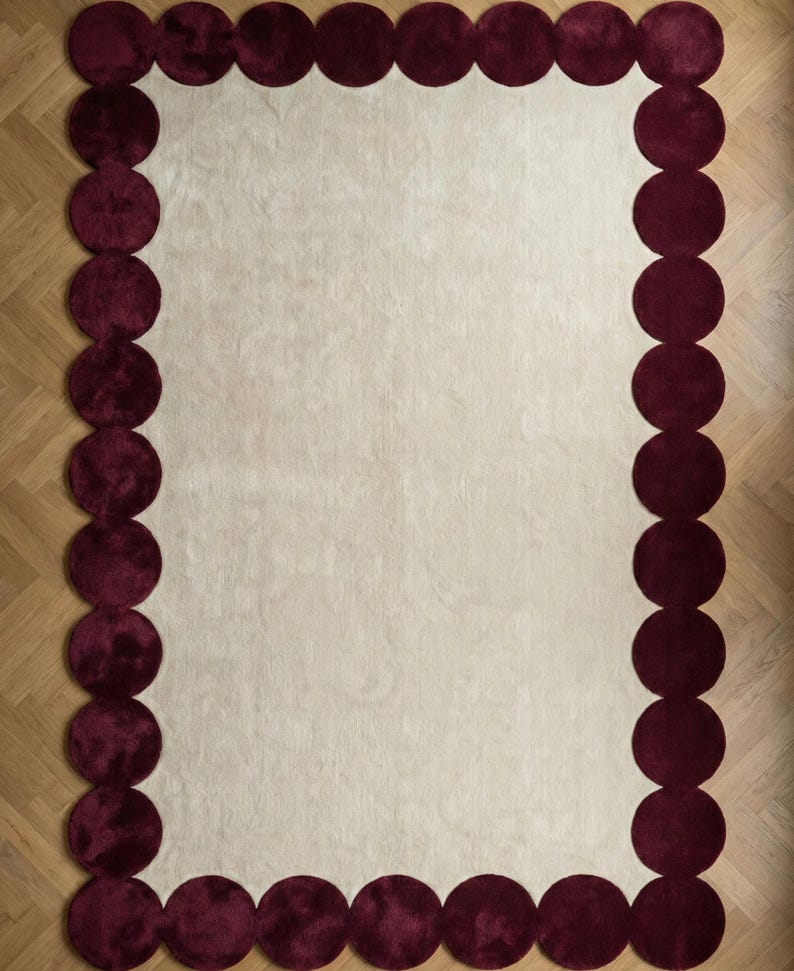 Hand tufted wool rug size 6x9 designed with a unique burgundy bubble border and subtle ivory base. Positioned in a stylish lounge near plants and modern furniture, the plush surface enhances warmth and texture. Ideal for homeowners seeking a fashionable yet timeless living room upgrade.