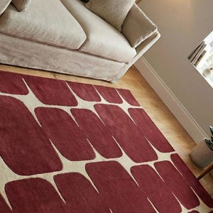 Handmade hand tufted wool rug featuring a contemporary geometric pattern in deep burgundy shades, placed in a bright living room with beige sofa, wooden chair, and natural light. This artisan crafted area rug offers a plush texture and premium quality, making it ideal for living room floor decor, bedroom styling, or modern office interiors. A perfect blend of modern design and cozy comfort.