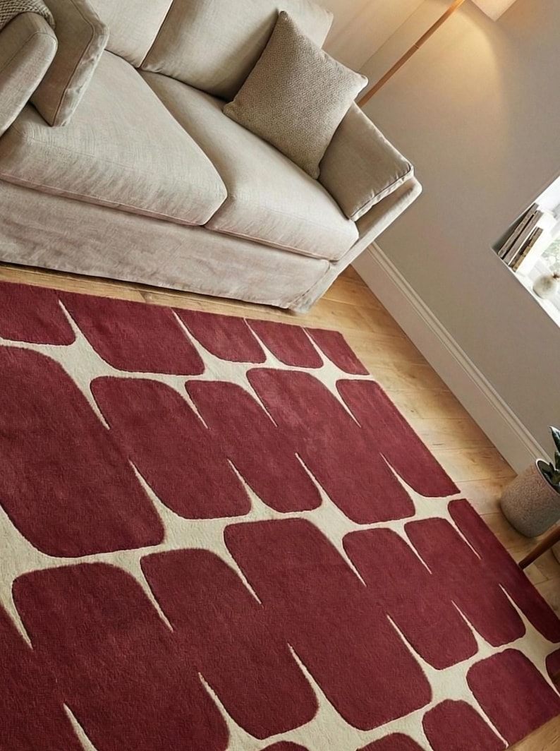 Handmade hand tufted wool rug featuring a contemporary geometric pattern in deep burgundy shades, placed in a bright living room with beige sofa, wooden chair, and natural light. This artisan crafted area rug offers a plush texture and premium quality, making it ideal for living room floor decor, bedroom styling, or modern office interiors. A perfect blend of modern design and cozy comfort.