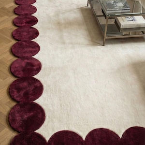 Modern ivory and burgundy hand tufted wool rug in size 9x12 featuring artistic rounded scallop borders surrounding a calm neutral field. Styled beneath a sofa in an elegant apartment, the rug defines the seating area and adds designer appeal. Thick pile, soft finish and handcrafted quality make it perfect for upscale living room decor.