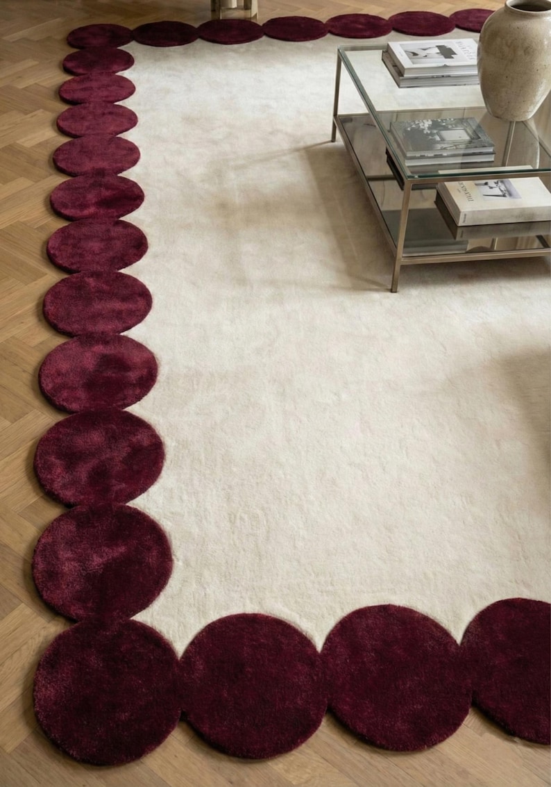 Modern ivory and burgundy hand tufted wool rug in size 9x12 featuring artistic rounded scallop borders surrounding a calm neutral field. Styled beneath a sofa in an elegant apartment, the rug defines the seating area and adds designer appeal. Thick pile, soft finish and handcrafted quality make it perfect for upscale living room decor.