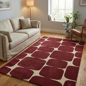 Hand tufted burgundy geometric rug displayed in a modern living room with wooden flooring, beige sofa, floor lamp, indoor plants, and natural sunlight coming through a large window. This handmade wool area rug features a bold abstract geometric pattern in deep burgundy and ivory tones, adding warmth, luxury, and contemporary style to living room, bedroom, or office home decor. Perfect modern handmade rug for stylish interiors.