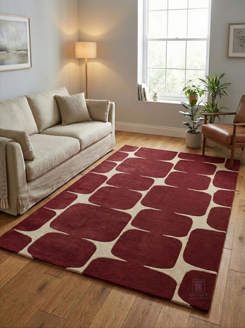 Hand tufted burgundy geometric rug displayed in a modern living room with wooden flooring, beige sofa, floor lamp, indoor plants, and natural sunlight coming through a large window. This handmade wool area rug features a bold abstract geometric pattern in deep burgundy and ivory tones, adding warmth, luxury, and contemporary style to living room, bedroom, or office home decor. Perfect modern handmade rug for stylish interiors.