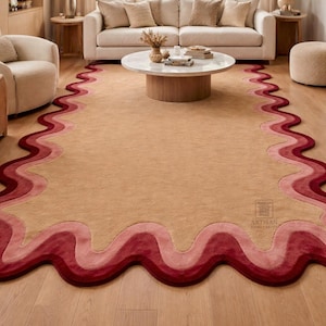 Op de afbeelding: Een beige vloerkleed met een golvende, gelaagde rand in roodtinten. Het kleed ligt op een houten vloer in een woonkamer, met een bank, stoelen en een salontafel op de achtergrond.