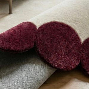 Luxury scalloped area rug 8x10 with deep burgundy circular trim and creamy center, hand tufted from premium wool. Displayed in a minimalist living space with wooden flooring and natural daylight, this carpet offers comfort underfoot while acting as a striking statement piece for modern and contemporary interiors.