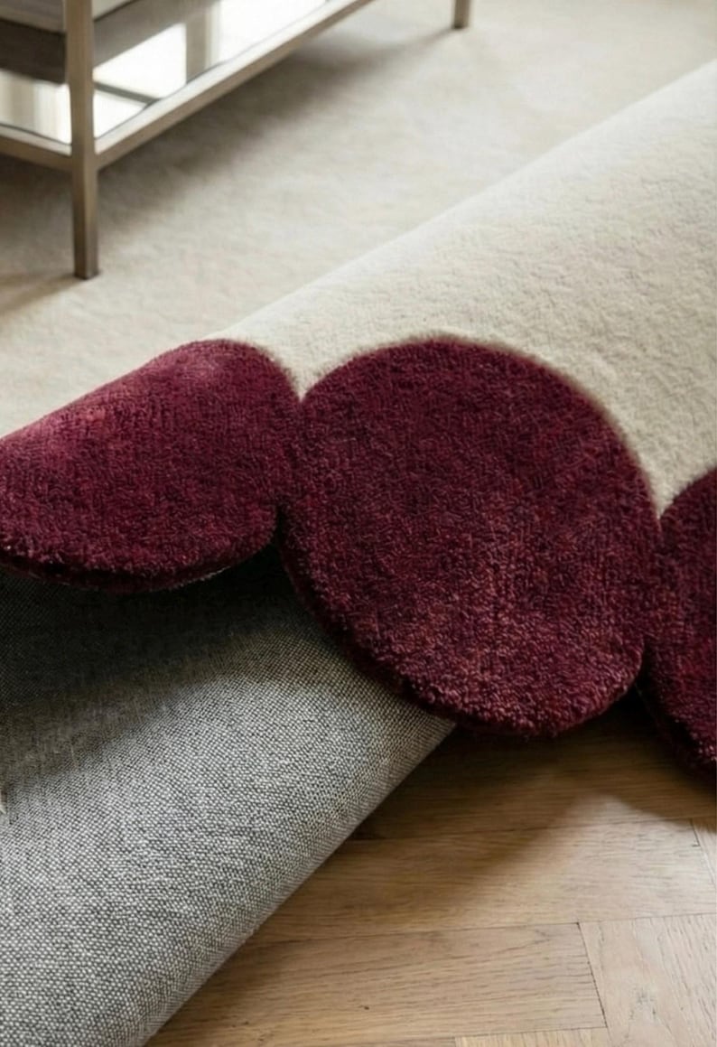 Luxury scalloped area rug 8x10 with deep burgundy circular trim and creamy center, hand tufted from premium wool. Displayed in a minimalist living space with wooden flooring and natural daylight, this carpet offers comfort underfoot while acting as a striking statement piece for modern and contemporary interiors.