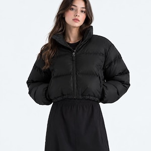 May include: A black cropped puffer jacket featuring a high collar, long sleeves with elastic cuffs, and a front zipper closure. The jacket has a quilted design with horizontal baffles and is worn over a black top and a black skirt.
