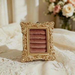 May include: A rectangular jewelry display stand featuring an ornate gold-colored frame with intricate scrollwork and floral detailing. The interior contains five horizontal rows of soft, dusty rose-colored velvet padding designed to hold rings.