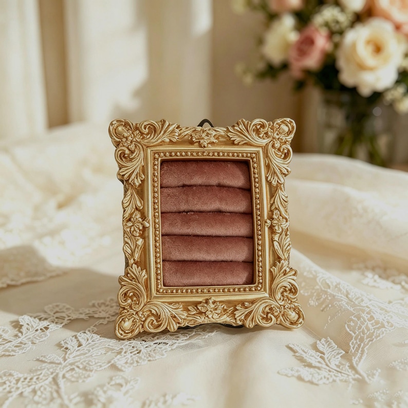 May include: A rectangular jewelry display stand featuring an ornate gold-colored frame with intricate scrollwork and floral detailing. The interior contains five horizontal rows of soft, dusty rose-colored velvet padding designed to hold rings.