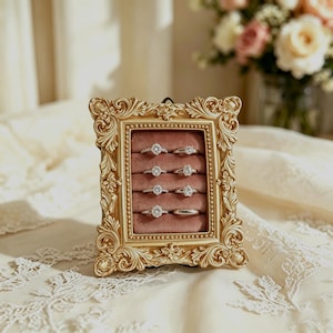 May include: A rectangular gold-toned ornate picture frame repurposed as a ring display stand. The interior features a dusty rose velvet cushion with four horizontal slots, each holding two silver-toned rings with clear stones and smooth bands.