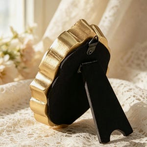 May include: A small, oval-shaped picture frame with a metallic gold finish and a scalloped edge. The back of the frame is covered in black velvet material and features a black easel-style stand and a small metal hook for wall mounting.