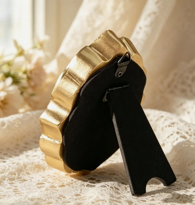 May include: A small, oval-shaped picture frame with a metallic gold finish and a scalloped edge. The back of the frame is covered in black velvet material and features a black easel-style stand and a small metal hook for wall mounting.