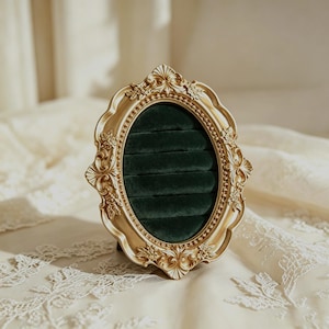 May include: An oval ring display holder featuring an ornate gold-colored frame with intricate floral and scrollwork detailing. The interior is lined with dark green velvet fabric, containing five horizontal slots designed for holding rings.