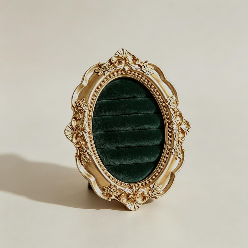 May include: An oval ring display holder featuring an ornate gold-colored frame with intricate floral and scrollwork detailing. The interior is lined with dark green velvet fabric, containing five horizontal slots designed to hold rings securely.