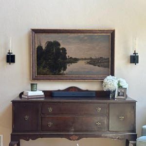 May include: A dark wood sideboard with two cabinets and two drawers, topped with a landscape painting in a dark frame, a soundbar, a stack of books, a candle, a vase of white hydrangeas, and a small framed photograph, flanked by two wall sconces.