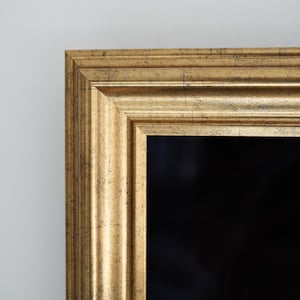 Könnte beinhalten: Nahaufnahme eines goldfarbenen Bilderrahmens mit schwarzem Innenraum. Der Rahmen hat ein detailliertes, strukturiertes Design und eine leicht verwitterte Oberfläche. Der Rahmen ist vor einer schlichten, hellfarbigen Wand angebracht.
