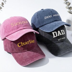 May include: Three baseball caps in shades of pink, blue, and black are stacked. The pink cap has "Sienna" embroidered in gold, the burgundy cap has "Charlie", the blue cap has "DOG MOM", and the black cap has "DAD EST. 2026".