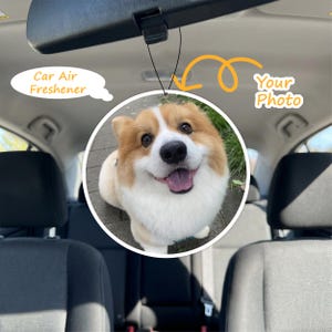 May include: A circular car air freshener hanging from a rearview mirror, featuring a custom printed photograph of a fluffy tan and white Corgi dog with a happy expression, tongue out, and dark eyes, suspended by a thin black elastic cord.