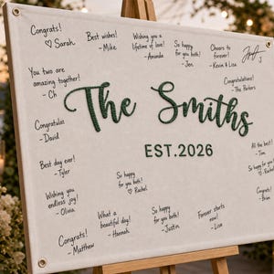 May include: A rectangular white fabric guest book sign featuring the dark green embroidered text The Smiths and EST. 2026. The sign includes metal grommets in the corners and is filled with various handwritten congratulatory messages in black ink.