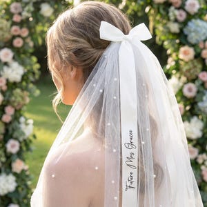 Pode incluir: Um véu de noiva branco com detalhes de pérolas e uma fita de cetim branca. A fita apresenta o texto "Future Mrs. Gracee" em fonte cursiva. O véu é preso com um laço branco.