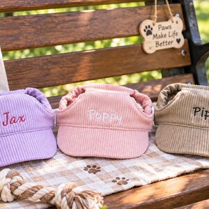 May include: Three corduroy pet visors in lavender, light pink, and tan. Each visor features an elastic strap and is embroidered with a name: Jax in red, Poppy in white, and Pip in black. The visors are designed for pets to wear on their heads.