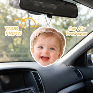 May include: A car air freshener with a smiling baby wearing a floral headband. Text reads "YOUR PHOTO" and "Car Air Freshener". The freshener is suspended from a car's rearview mirror, with the dashboard visible in the foreground.