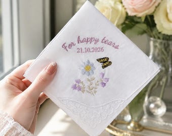 May include: A white square handkerchief featuring embroidered text that reads For happy tears and the date 21.10.2026 in purple thread. Below the text is an embroidered design of a light blue daisy, purple wildflowers, and a yellow butterfly.