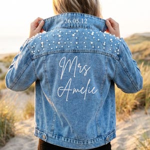 May include: A light blue denim jacket adorned with pearl detailing on the shoulders. The back displays the text "Mrs Amelie" in white script, with the date "26.05.12" above the collar. A fashionable and personalized bridal jacket.