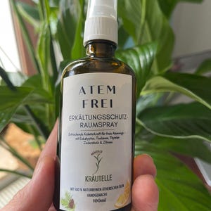 Könnte beinhalten: Eine braune Glas-Sprühflasche mit weißem Etikett mit der Aufschrift "ATEM FREI" und "ERKÄLTUNGSSCHUTZ-RAUMSPRAY". Das Etikett enthält auch den Text "KRÄUTELLE" und "100ml". Die Flasche wird vor einem grünen, belaubten Hintergrund gehalten.